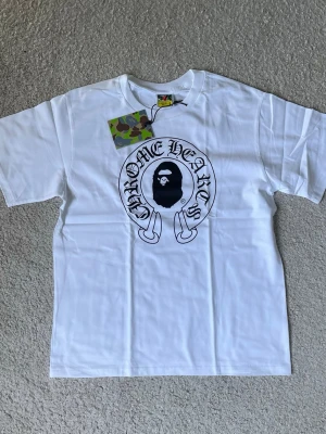 Vit BAPE x Chrome Hearts t-shirt - Säljer en vit t-shirt från BAPE med Chrome Hearts-tryck på bröstet och den klassiska BAPE-loggan på ärmen. T-shirten har rund hals och korta ärmar. Materialet är mjuk bomull och passformen är normal. Perfekt för dig som gillar streetwear och unika samarbeten.