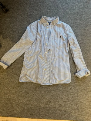 Blårandig skjorta från Polo Ralph Lauren - Klassisk blårandig skjorta från Polo Ralph Lauren med button-down krage och broderad logga på bröstet. Skjortan har långa ärmar, vita knappar och är tillverkad i bomull. Perfekt för en clean och stilren look. Är ca 175 och väger 65 och den är lite kort i armarna. 