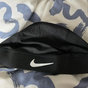 Nike svart Dri-FIT skull cap - Snygg svart skull cap från Nike med vit swoosh-logga framtill. Tillverkad i Dri-FIT-material som transporterar bort fukt och håller dig torr. Perfekt för träning eller sportaktiviteter där du vill ha en tight och bekväm passform.