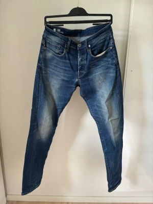 G star Straight tapered - Säljer ett par blå jeans med raka ben och snygga slitningar på framsidan. Jeansen har klassisk femficksmodell, knappgylf och kontrastsömmar. Materialet är denim i bomull och passformen är avslappnad. Perfekta för en casual look.