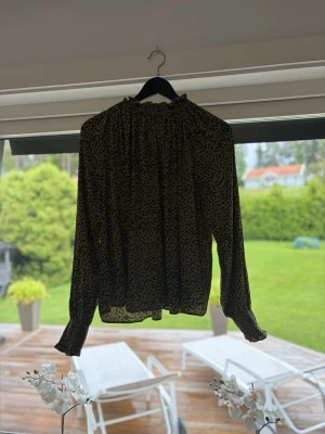 Grönmönstrad blus med volangkrage - Säljer en tunn, grön blus med svart småprickigt mönster och volangdetaljer vid halsen. Blusen har långa ärmar med resår och volang vid ärmslut. Perfekt för dig som gillar en luftig och trendig stil. Skulle säga att de är en S