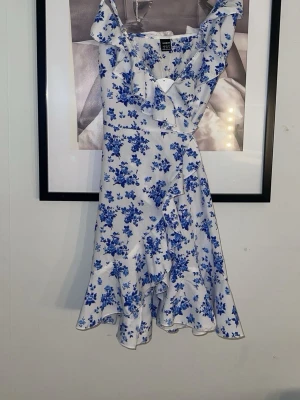 Vit klänning med blå blommor och volang - Säljer en söt vit kortklänning med blått blommönster och volangdetaljer. Klänningen har omlott-design, smala axelband. Perfekt för sommarens alla tillfällen! Djur finns i hemmet, tvättas innan leverans.💙