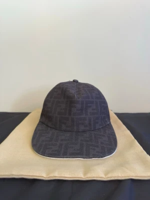 Svart keps med FF-Logga - Säljer en svart keps med det klassiska FF-monogrammet över hela kepsen. Kepsen har böjd skärm, justerbar rem bak och är tillverkad i ett tygmaterial. Perfekt för dig som vill ha en stilren accessoar med lyxig känsla.