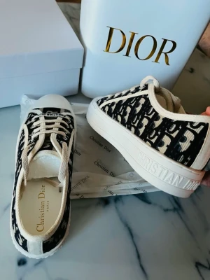 Dior Oblique sneakers i svart och vitt - Snygga Dior Oblique sneakers med marinblå och beiget monogrammönster, vita snören och robust vit sula med stjärndetalj undertill. Klassisk låg modell med 'Christian Dior' tryckt på sidan och insidan. Tillverkade i canvas och gummi för en trendig och exklusiv look.