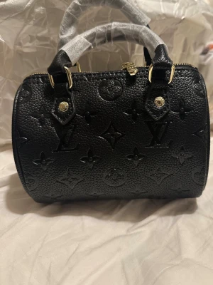 Svart liten handväska - Snygg svart handväska i präglat läder med det klassiska monogrammönstret. Väskan har guldfärgade metalldetaljer och två handtag. Perfekt storlek för det viktigaste och en ikonisk accessoar till din outfit.