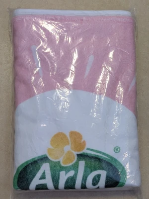Rosa och vit Arla-tryckt badlakan  - Säljer ett badlakan med tryck från Arla i rosa och vitt, med Arlas logga i grönt och gult. Badlakanet är vikt i förpackning. Perfekt för dig som gillar unika eller reklamrelaterade badlakan Oöppnad plast förpackning 