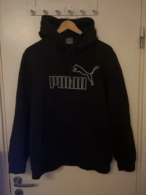 Svart hoodie från Puma med logga - Svart hoodie från Puma med stor vit logga och text på bröstet. Klassisk känguruficka framtill och justerbar huva med dragsko. Perfekt för en avslappnad och sportig stil.