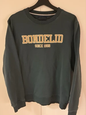 Sweatshirt Bondelid - Mörkgrön sweatshirt från Bondelid med tryckt text 'BONDELID SINCE 1958' i beige på bröstet. Tröjan har rund halsringning, ribbade muddar vid ärmslut och nederkant. Perfekt för en avslappnad och stilren look.