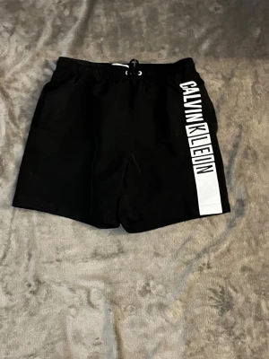 Svarta badbyxor Calvin Klein - Snygga svarta badbyxor från Calvin Klein med vit logga längs ena benet. Klassisk modell med resår och dragsko i midjan samt fickor på sidorna och bak. Tillverkade i snabbtorkande material, perfekta för stranden eller poolen. Vid frågor hör av er priset kan alltid diskuteras 