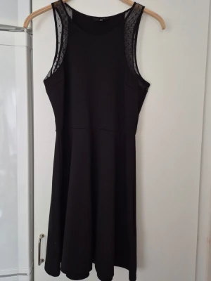 Klänning - Snygg svart klänning med meshdetaljer vid axlarna och ärmlös design. Klänningen har en rund halsringning och en lätt utställd kjol som ger en feminin siluett. Perfekt för dig som gillar stilrena plagg med en twist.