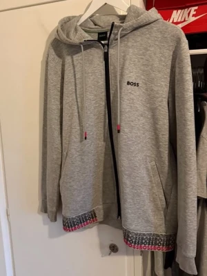 Grå zip hoodie från BOSS - Snygg grå zip hoodie från BOSS med huva och dragsko. Diskret BOSS-logga på bröstet och färgglad detalj med BOSS-tryck längs nederkanten. Mjukt material och två fickor framtill. Perfekt för en avslappnad och trendig look. Storlek L men passar även m. Priset kan diskuteras !!