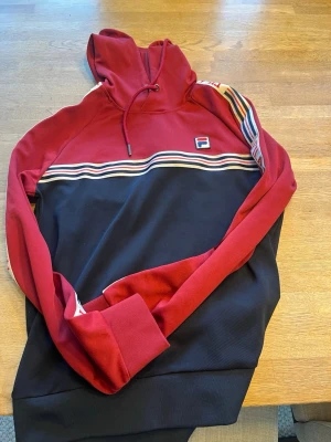 Röd och svart hoodie från FILA - Snygg hoodie från FILA i rött och svart med klassiska ränder över bröstet och ärmarna. Hoodien har dragsko i huvan och en broderad FILA-logga på bröstet. Tillverkad i mjuk polyester för en bekväm känsla. Perfekt för en sportig och avslappnad stil.