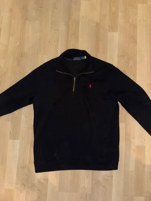 Svart stickad tröja från Polo Ralph Lauren - Svart stickad tröja från Polo Ralph Lauren med halv dragkedja och klassisk röd logga broderad på bröstet. Tröjan har hög krage och långa ärmar, perfekt för lager-på-lager-stil. Materialet är mjukt och bekvämt.