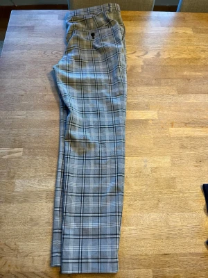 Rutiga kostymbyxor i grått - Snygga kostymbyxor med klassiskt rutmönster i grått, svart och vitt. Byxorna har raka ben och en bakficka med knapp. Perfekta för en stilren och trendig look. Materialet känns mjukt och följsamt.