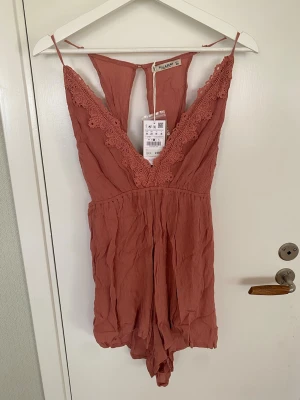 Rostfärgad spetsbody från Pull&Bear - Supersnygg rostfärgad body från Pull&Bear med tunna axelband och djup V-ringning dekorerad med spetsdetaljer. Tillverkad i ett mjukt, lätt skrynklat tyg som känns bekvämt mot huden. Perfekt för dig som vill ha något både trendigt och bekvämt.