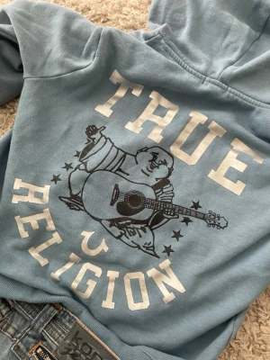 Blå hoodie från True Religion - Snygg ljusblå hoodie från True Religion med stor grafisk print på ryggen av en Buddha med gitarr och texten 'True Religion'. Klassisk känguruficka framtill, dragsko i huvan och liten logga på bröstet. Perfekt för en avslappnad och trendig stil.