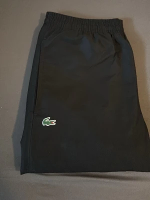 Svarta mjukisbyxor från Lacoste - Svarta mjukisbyxor från Lacoste med klassisk grön krokodil-logga på benet. Byxorna har resår i midjan och är tillverkade i ett mjukt och bekvämt material, perfekta för chill dagar. Snygg och enkel design som funkar till det mesta.
