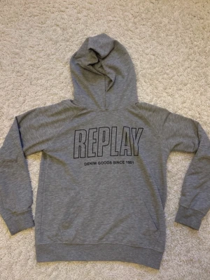Grå hoodie från Replay - Snygg grå hoodie från Replay med stor logga och texten 'DENIM GOODS SINCE 1981' på bröstet. Klassisk modell med huva och ribbade muddar vid ärmslut och nederkant. Perfekt för en avslappnad och trendig look. Kan även passa in till en grisch och rik look. Vid minsta fråga skriv och priset kan alltid diskuteras 