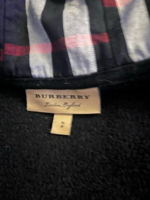 Svart hoodie från Burberry med dragkedja - Snygg svart hoodie från Burberry med dragkedja framtill och klassiskt rutigt foder i huvan. Hoodien har fickor på magen och justerbar dragsko i huvan. Perfekt för dig som vill ha en stilren och exklusiv look med en touch av Burberrys ikoniska mönster.