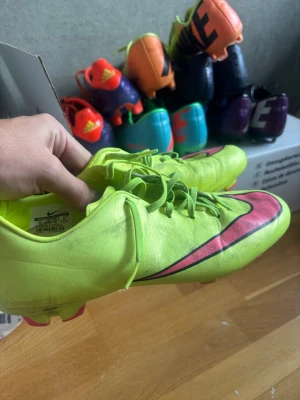 Nike Mercurial neon-gula fotbollsskor - Säljer ett par Nike Mercurial fotbollsskor i neon-gult med rosa detaljer och klassisk Nike-swoosh på sidan. Skorna har snörning och är tillverkade i ett lätt syntetmaterial för bra bollkontroll och snabbhet på planen. Perfekta för dig som vill sticka ut på fotbollsplanen.