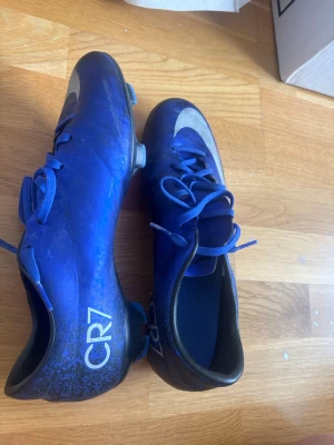 Blå Nike CR7 fotbollsskor - Säljer ett par blå Nike CR7 fotbollsskor med silverfärgad swoosh och CR7-logga på hälen. Skorna har snörning och är tillverkade i syntetmaterial för bra grepp på planen. Perfekta för dig som vill sticka ut på matchen!