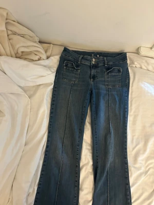 Blå bootcut jeans med fickdetaljer - Säljer ett par blå bootcut jeans med dubbla knappar i midjan och stora fyrkantiga framfickor. Jeansen har markerad söm längs benen och breda hällor för bälte. Klassisk tvättad denim-look och raka ben med lätt utsvängning.