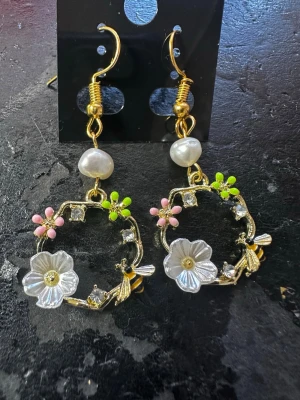 Blommiga örhängen med pärldetaljer - Unika örhängen i guldton med vita pärlor, dekorerade med små rosa och gröna blommor samt vita blommor i plast. Runda hängen med gnistrande stenar och detaljer i metall. Perfekta för dig som gillar färgglada accessoarer med blommig touch.