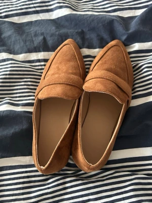 Bruna mockaloafers med spetsig tå - Oanvända loafers i brun mocka med spetsig tå och klassisk rem över foten. Skorna har låg klack och en stilren design som passar till många olika outfits. Perfekt för dig som gillar en trendig och enkel look.