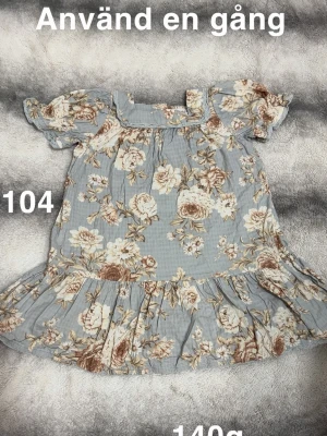 Blommig kortklänning med volang - Söt kortklänning i ljusblått med beige och bruna blommor. Klänningen har volang nertill, korta puffärmar och en söt krage. Perfekt för sommarens varma dagar.🌸Använd en gång. Storlek 104