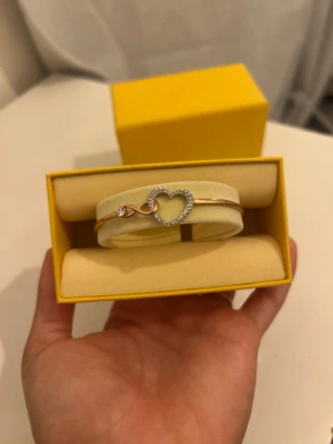 Swarovski Guldarmband med hjärta och stenar - Snyggt armband i guldton med ett glittrande hjärta dekorerat med små klara stenar. Armbandet har en elegant och tunn design med en liten steninfattning vid låset. Perfekt accessoar för att lyfta din stil.