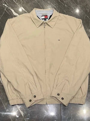 Beige overshirt från Tommy Hilfiger - Klassisk beige vindjacka från Tommy Hilfiger med dragkedja framtill och liten logga på bröstet. Storlek XL men sitter som en L. Jackan har krage, långa ärmar med knappar vid mudden och elastiska ärmslut. Perfekt för dig som gillar stilren och enkel design.
