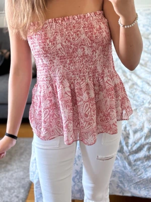Rosa och vit smockad peplum topp - Söt peplumtopp i rosa och vitt med smockad överdel och volangig nederkant. Toppen har ett paisleyinspirerat mönster och är axelbandslös, perfekt för varma dagar. Lätt och luftig känsla, passar snyggt till jeans eller kjol. Använd fåtal gånger. Från gina tricot. Passar S och M! Inga fläckar!