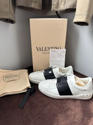 Valentino Garavani vita sneakers - Snygga vita sneakers från Valentino Garavani med bred svart läderdetalj över snörningen och diskret logga på plösen. Klassisk låg modell med vita skosnören och gummisula. Perfekta för dig som vill ha en clean men lyxig look.