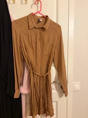 Beige skjortklänning med bälte H&M - Stilren beige skjortklänning från H&M med knappar framtill, lång ärm och ett matchande bälte i midjan. Klänningen har klassisk krage och en bröstficka för extra detalj. Perfekt för dig som gillar enkel och avslappnad stil. Strl 38 men passar 36