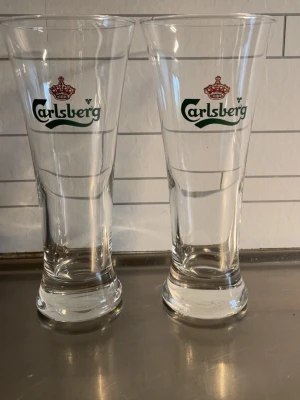 Två Carlsberg ölglas 80kr st - Säljer två höga ölglas från Carlsberg med klassisk logga i grönt och rött tryck. Glasen är genomskinliga och har en avsmalnande form som breddas upptill. Perfekta för att servera öl eller andra drycker.