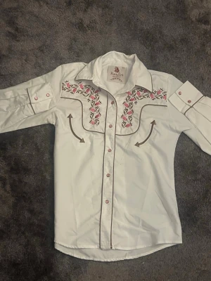 Vit westernskjorta med broderi - Vit westernskjorta från Ranger's med broderade rosa och gröna blommor på bröstet. Skjortan har långa ärmar, knappar framtill och dekorativa sömmar. Perfekt för dig som gillar cowboystil och unika detaljer.