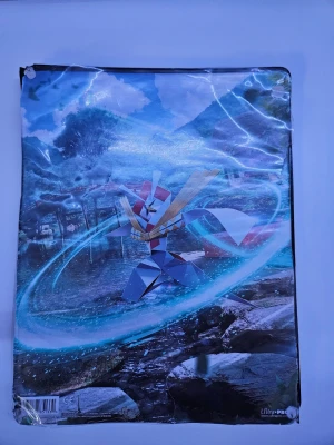 Pokémon pärm Ultra PRO motiv - Säljer en pärm med Pokémonmotiv från Ultra PRO, perfekt för att förvara samlarkort eller andra papper. Framsidan har en färgglad illustration av en Pokémon i en naturmiljö och baksidan är enfärgad svart. Materialet är plast med glansig yta.