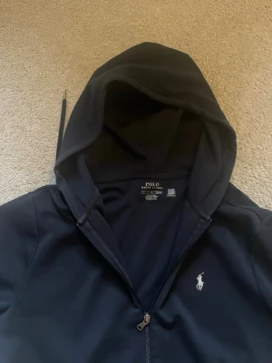 Svart hoodie från Polo Ralph Lauren - Snygg svart hoodie från Polo Ralph Lauren med dragkedja framtill och klassisk vit logga på bröstet. Hoodien har huva med dragsko och en stor magficka. Tillverkad i mjukt bomullsmaterial som är skönt att ha på sig. Perfekt för en chill och stilren look.