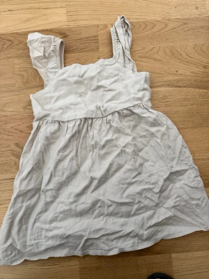 Ljusbeige ärmlös kortklänning i linne till barn  - Söt ljusbeige kortklänning i linne med breda axelband och rynkdetaljer upptill. Klänningen har en avslappnad passform och är perfekt för varma sommardagar. Enkel och stilren design som passar till många tillfällen. Storlek 98/10