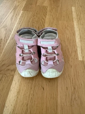 Rosa sportiga sandaler med kardborre - Säljer ett par rosa sandaler med sportig design och kardborreband. Sandalerna har meshdetaljer för extra ventilation och vita detaljer på sidorna samt tåskydd. Perfekta för varma dagar och aktiva äventyr.