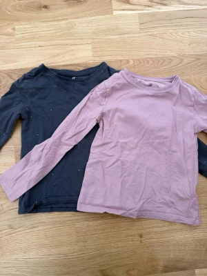 Två långärmade toppar från H&M - Säljer två basic långärmade toppar från H&M i storlek 110/116. En topp är ljusrosa och den andra är mörkgrå. Båda har rund halsringning och är tillverkade i mjuk bomull, perfekta för lager på lager eller som de är.