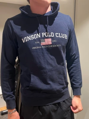 Marinblå hoodie Vinson Polo Club - Snygg marinblå hoodie från Vinson Polo Club med tryck framtill och amerikansk flagga. Klassisk känguruficka och justerbar huva med dragsko. Tillverkad i mjukt bomullsmaterial, perfekt för en avslappnad och sportig stil.
