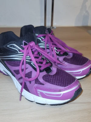 Lila och svarta sneakers med meshdetaljer - Snygga sneakers i lila och svart med vita och silvriga detaljer. Ovandelen har meshpartier för bra ventilation och skorna har matchande lila skosnören. Sulan är vit med svarta inslag och ger en sportig look. Perfekta för dig som gillar färg och komfort.