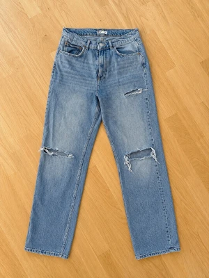 Blå mom jeans med slitningar - Säljer ett par ljusblå mom jeans med raka ben och slitna detaljer vid knäna och fickorna. Jeansen har klassisk femficksmodell, hög midja och är tillverkade i kraftigt denimtyg. Perfekta för en avslappnad och trendig look.
