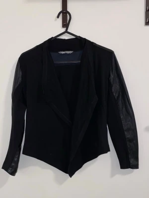 Svart skinnjacka med draperad front - Snygg svart skinnjacka med draperad öppen front och långa ärmar i skinn. Jackan har en cool mix av material med skinn på ärmar och axlar samt tyg på resten av jackan. Perfekt för en edgy look och passar till många olika stilar.