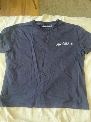 Mörkblå t-shirt med tryck - Säljer en mörkblå t-shirt med vit text 'MA CHÉRIE' på bröstet och stort tryck med hjärtan och text på ryggen. Klassisk passform och rund hals. Perfekt för dig som gillar enkla men snygga statement-plagg.