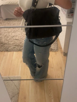 Svart axelväska med silverdetaljer - Snygg svart axelväska i tyg med coola silverfärgade metalldetaljer och justerbar axelrem. Väskan har en mjuk, lite slouchy form och flera fack på insidan för extra förvaring. Perfekt för dig som vill ha en stilren men ändå trendig väska till vardags.