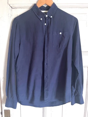 Mörkblå skjorta från Norse Projects - Klassisk mörkblå skjorta från Norse Projects med button-down krage och bröstficka med vit knapp. Skjortan har långa ärmar och stängs med knappar framtill. Tillverkad i mjuk bomull, perfekt för en clean och stilren look.
