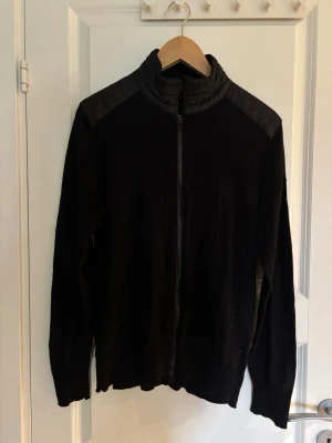 Svart ziptröja från Belstaff M/S - Snygg svart ziptröja från Belstaff med hög krage och quiltade axelpartier. Tröjan har hel dragkedja framtill och är tillverkad i Italien. Tröjan är något krympt, skulle säga att den passar bättre S. Hör gärna av er vid frågor eller önskemål om fler bilder.