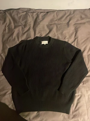 Svart stickad tröja Maison Margiela - Säljer en svart stickad tröja från Maison Margiela med ribbad struktur och rund hals. Tröjan har långa ärmar och de ikoniska vita sömmarna i nacken. Perfekt för dig som gillar minimalistisk och stilren design.